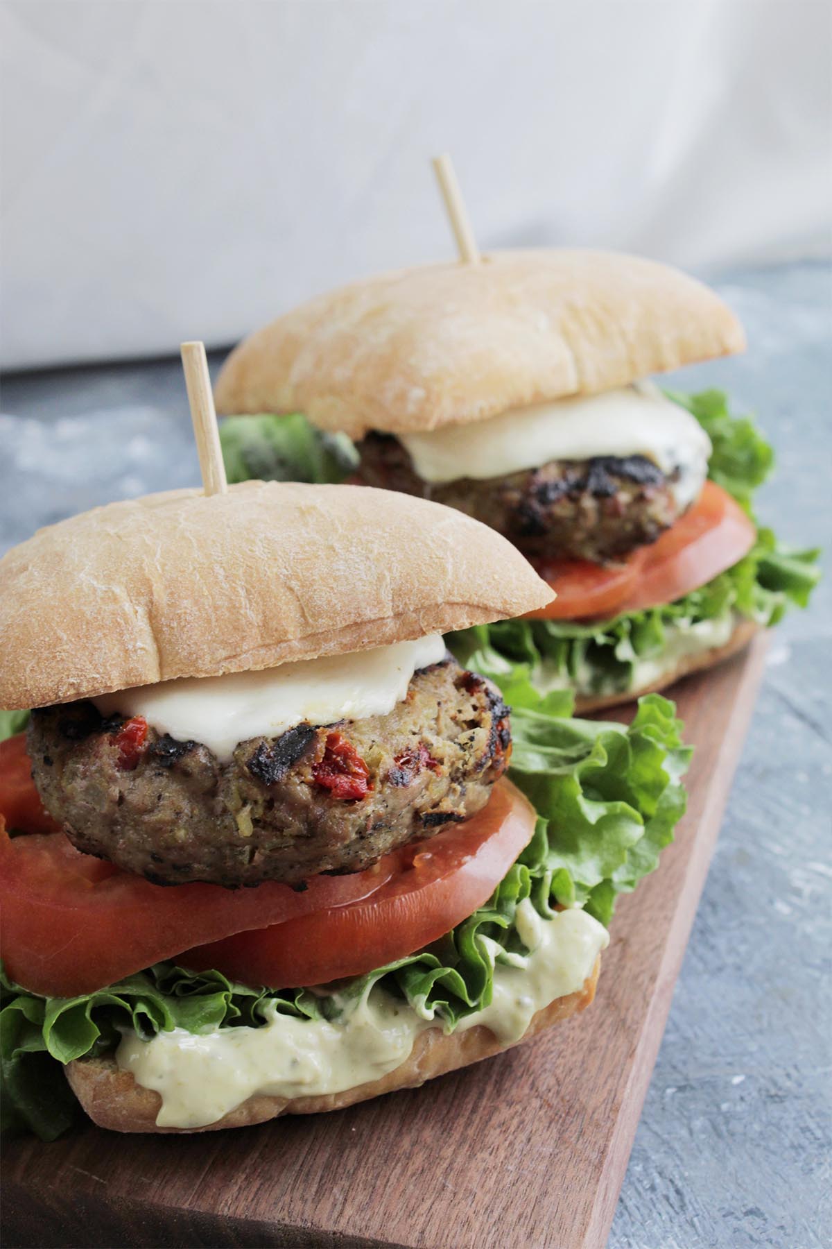 two burgers on ciabatta rolls sitting on wooden cutting board.