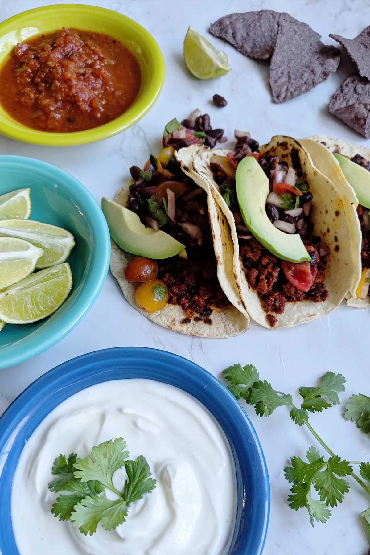 three street tacos surrounded by colorful bowls filled with toppings.