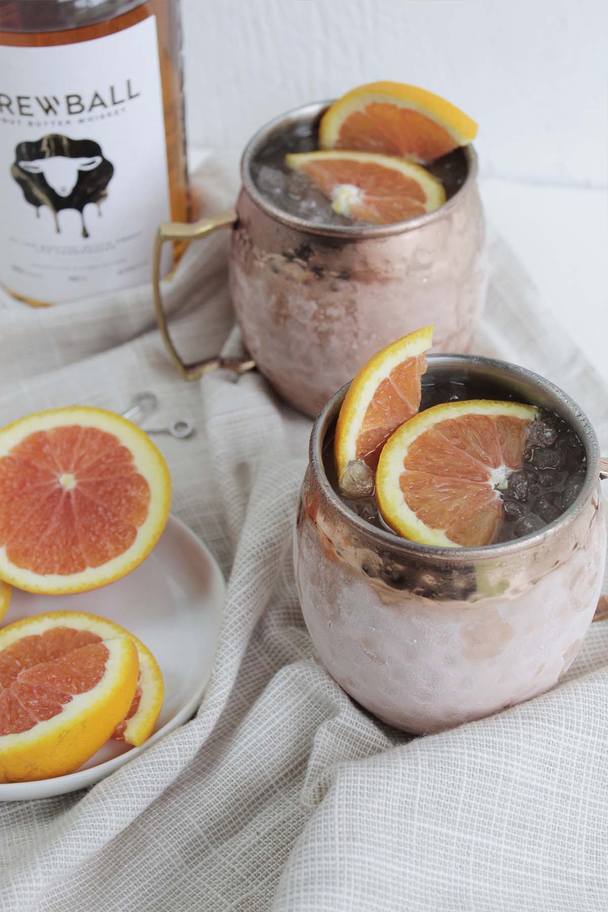 two copper cocktail mugs next to Skrewball bottle and orange slices.