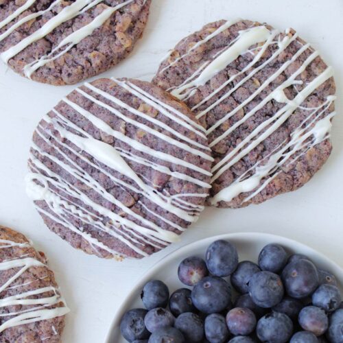 blueberry cookies topped with white chocolate drizzle next to a bowl of fresh blueberries.