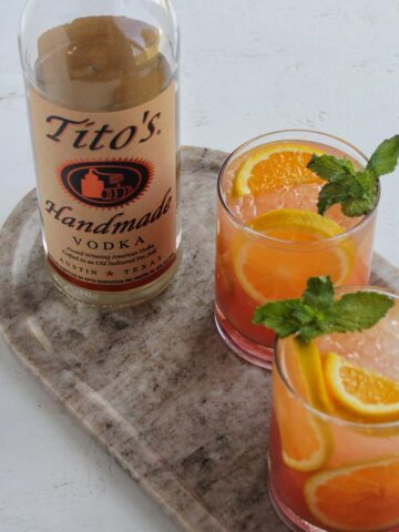two orange flavored cocktails next to Tito's vodka bottle.