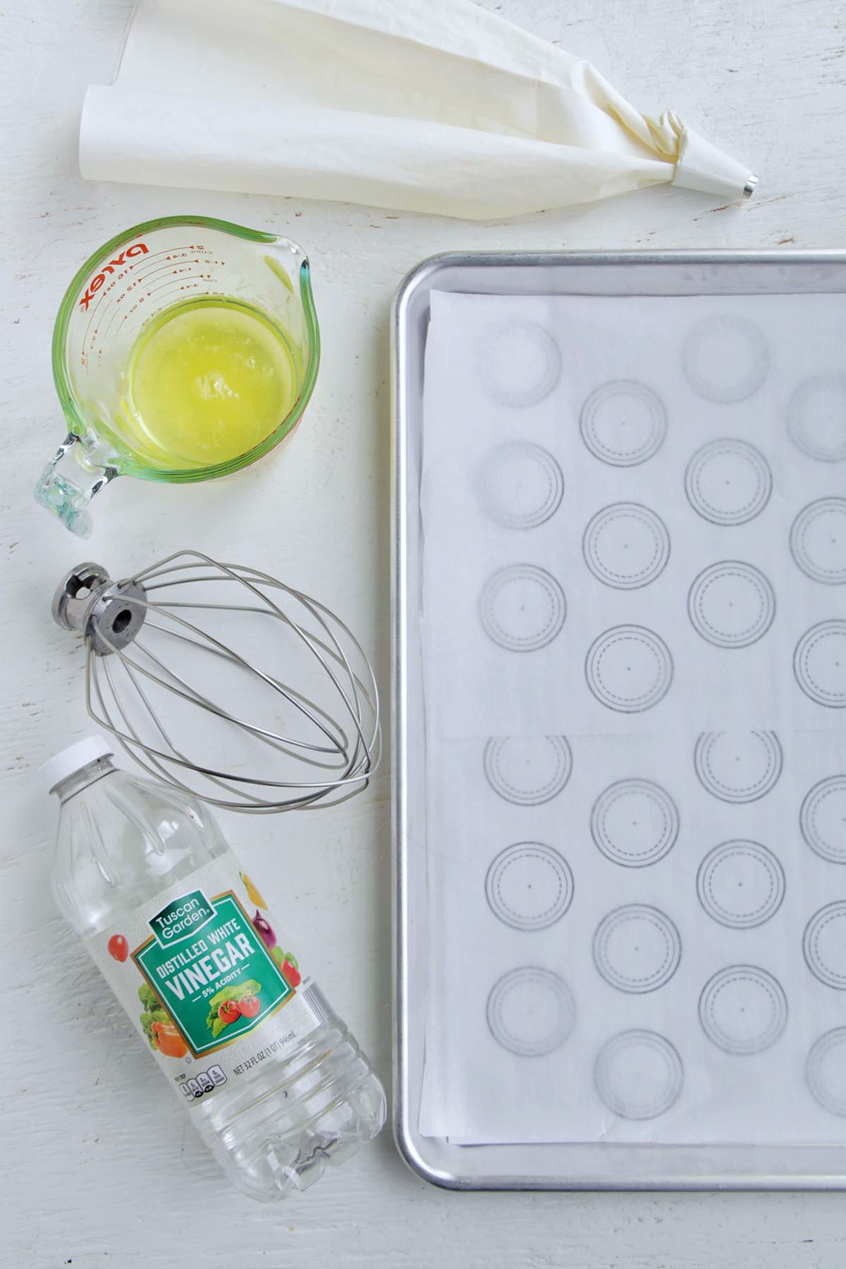 baking sheet with macaron template next to vinegar, whisk attachment, eggs, and piping bag.