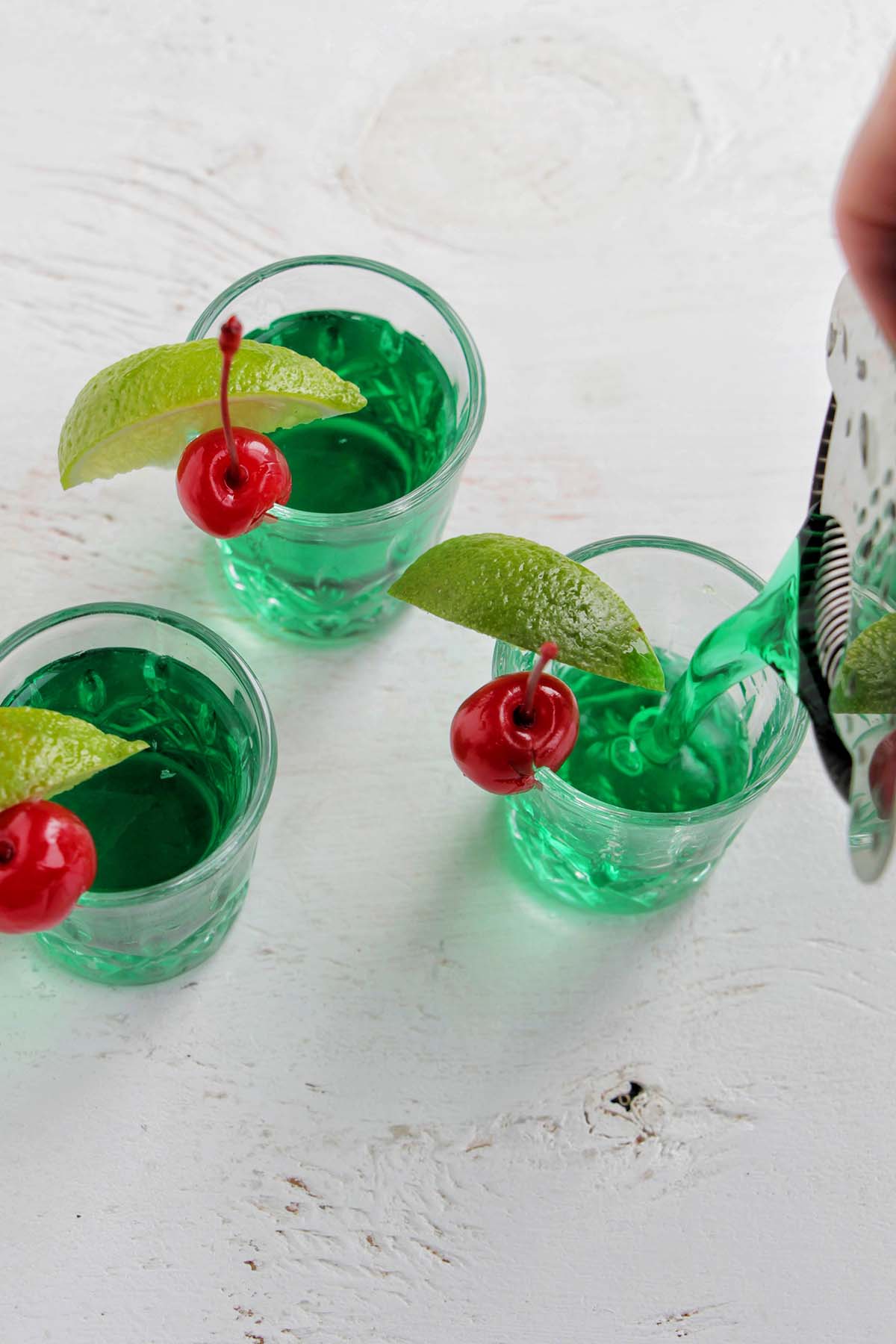 pouring three green shots garnished with a lime and cherry.