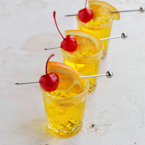 three yellow shots garnished with an orange and cherry.